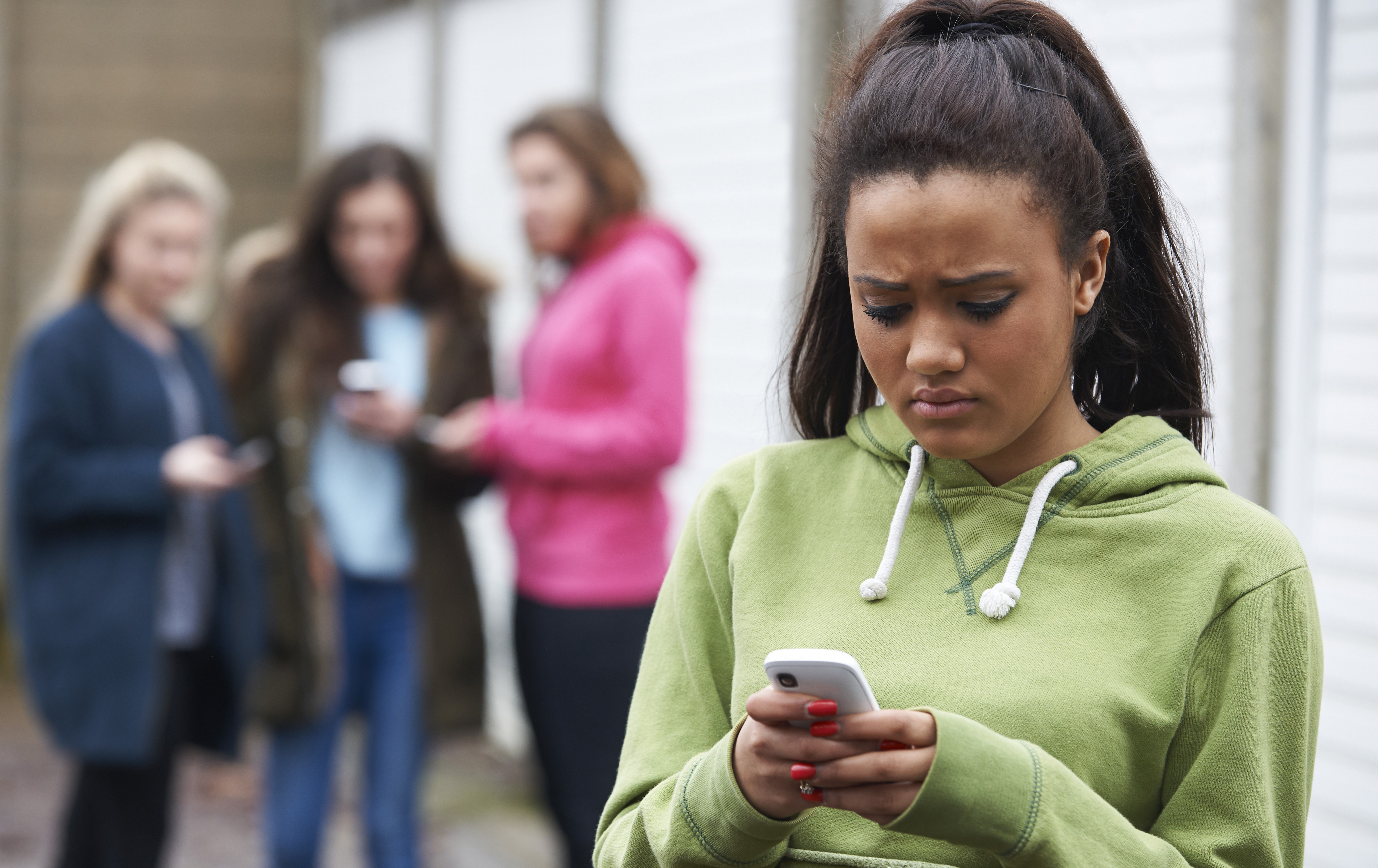 Meisje in groene trui kijkt bang naar haar smartphone terwijl andere meisjes in de achtergrond lachen