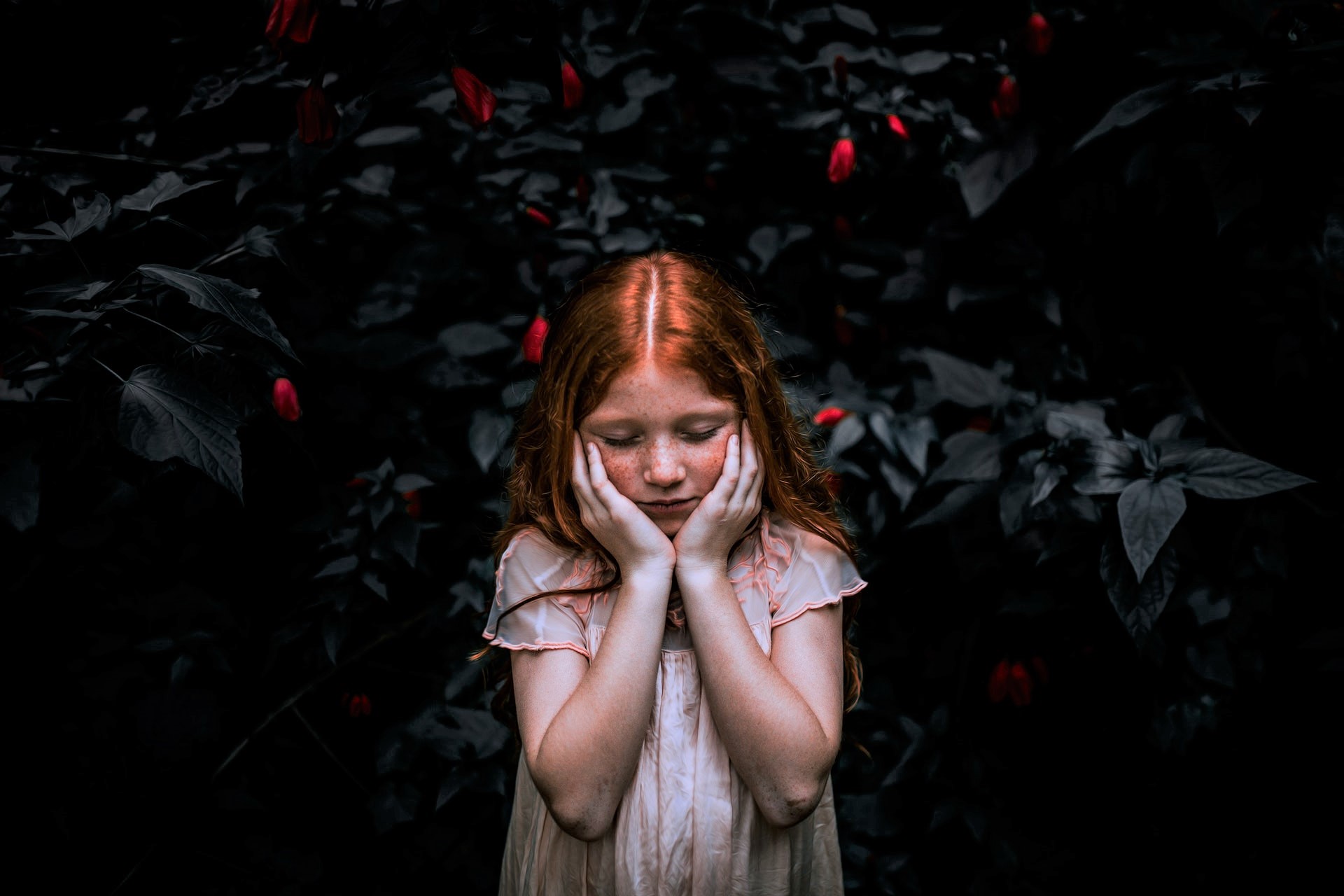 Ros meisje drukt handen tegen wangen tegen achtergrond van donkere struik