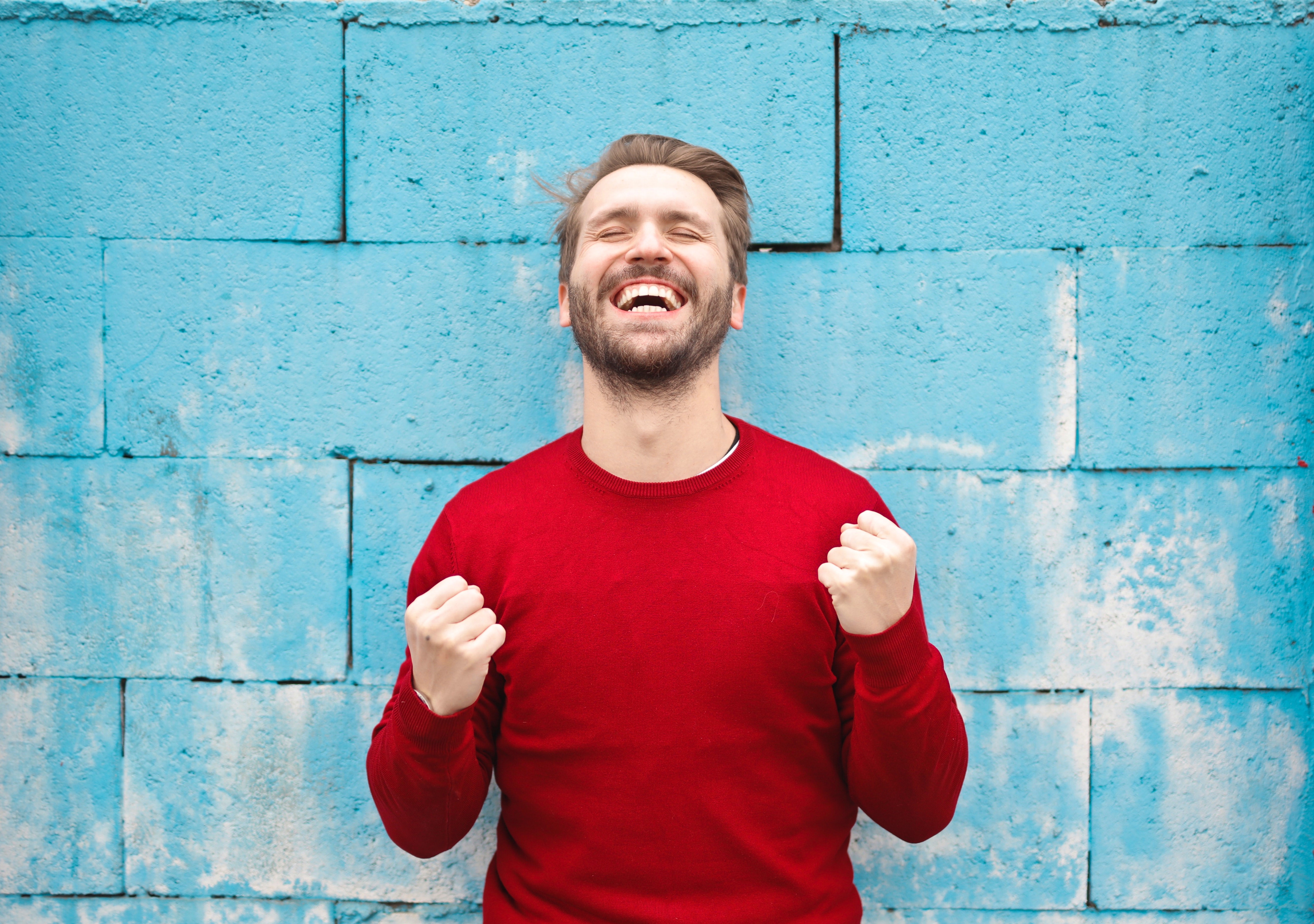 Man in rode trui tegen blauwe muur maakt tevreden yes-gebaar en lacht