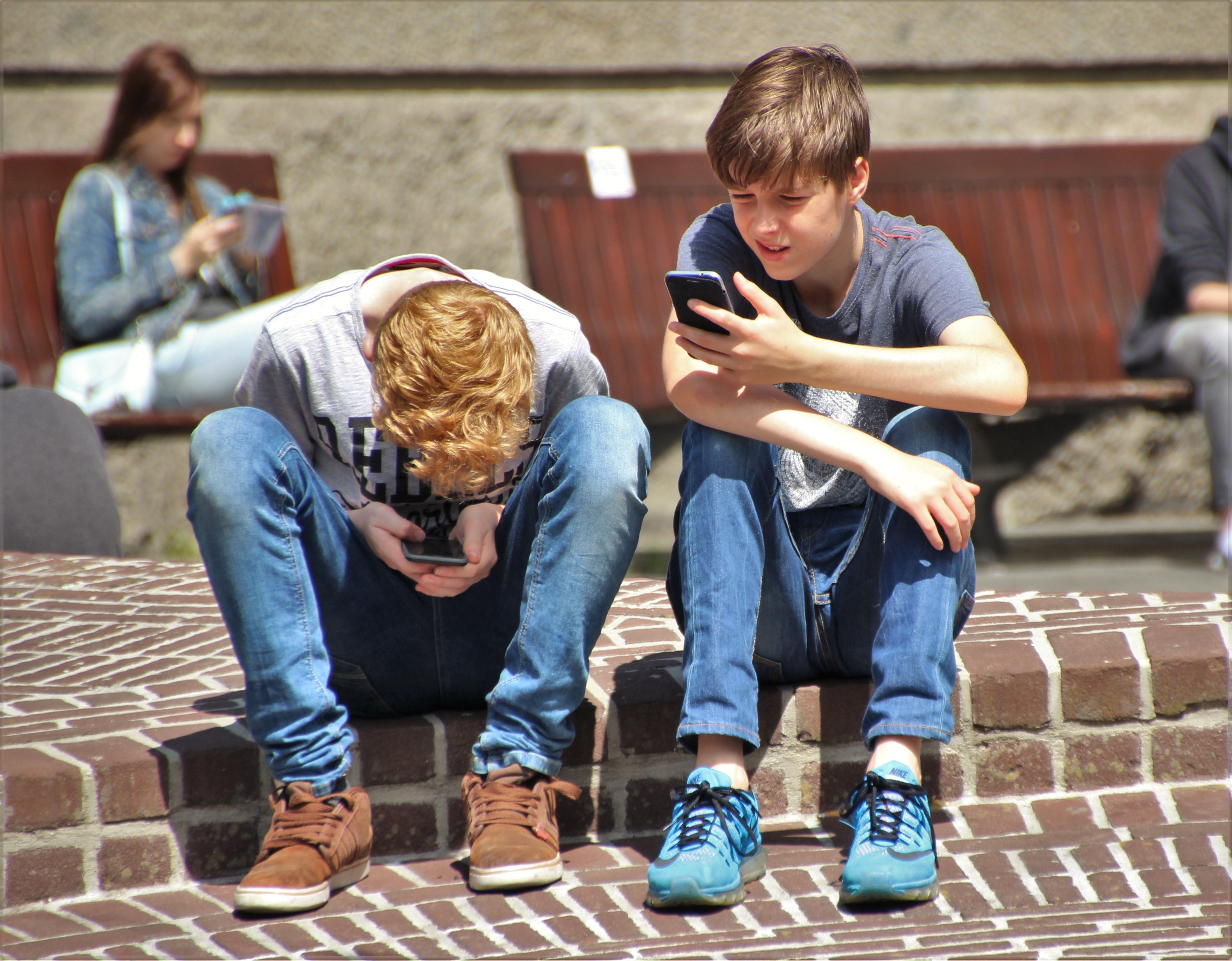 2 jongens zitten op pleintje, gebogen over hun smartphones