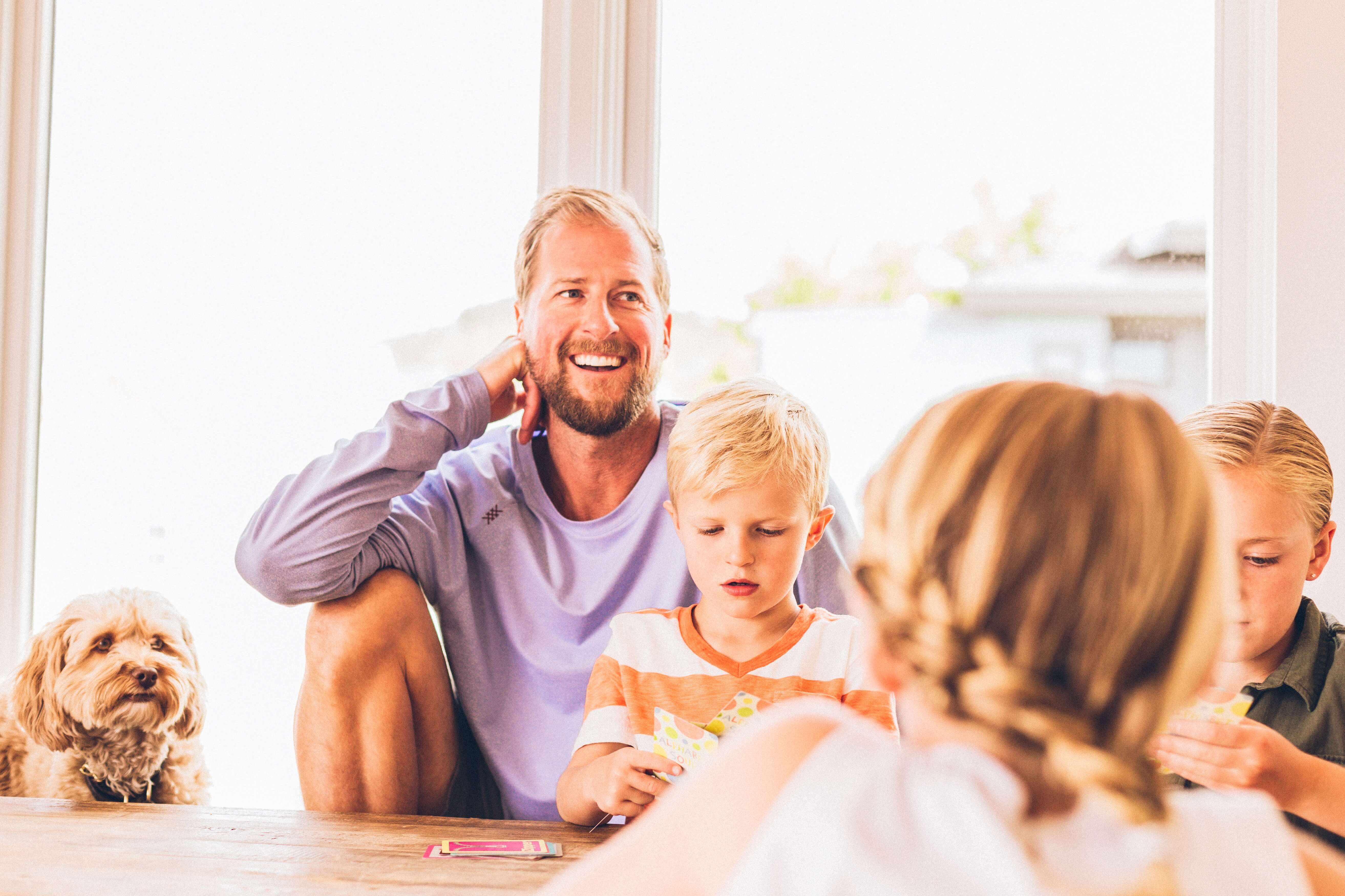 Gezin van 3 kinderen aan tafel, vader in paarse trui en hondje naast zich