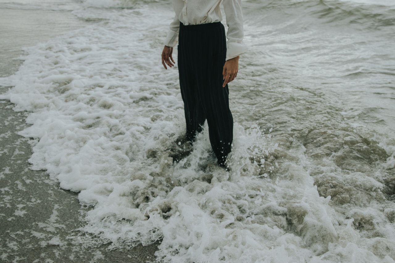 Persoon in zwarte broek staat met voeten in aanrollende golven op het strand